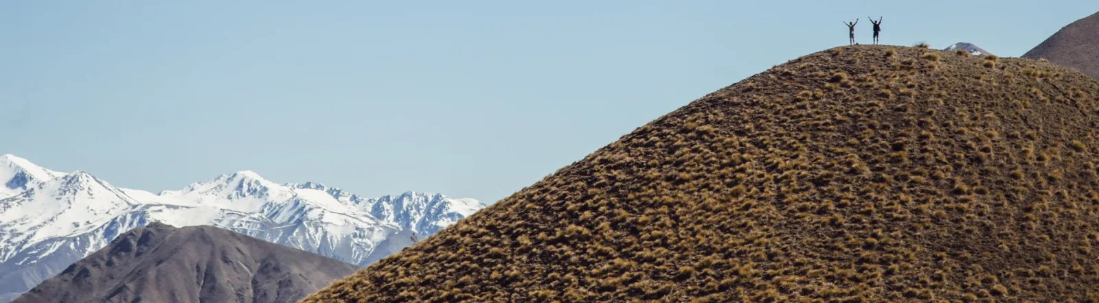 Two people on the top of a hill, with snowy mountains in the background