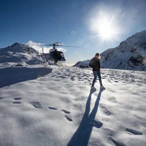Person on a snowing mountain walking away from a helicopter that is just behind them